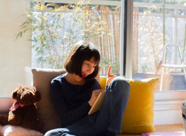 Mutlu çocuk gülüyor, şaşırtıyor, masa oyunlarında oynuyor, oyuncak ayıyla covid-19 kilit altında, güneşli dairede yeşil balkonlu yastıklara yaslanıyor.