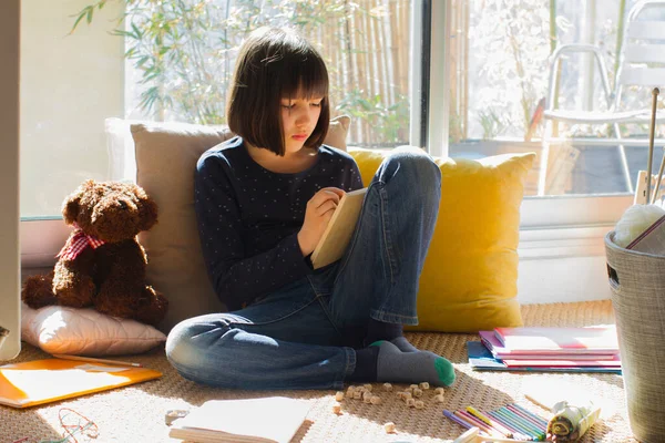 child playing with letters on board games studying for homeschooling with teddy bear, notebooks and pens on the floor - sunny home with green balcony