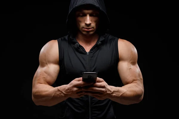 Muscular bodybuilder with phone in hands on a dark background. Sports nutrition. Bodybuilding nutrition supplements, sport, workout, healthy lifestyle concept.