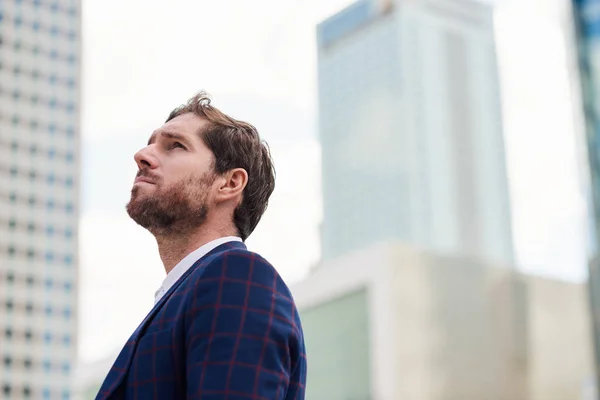 Ambitious young businessman wearing blazer looking at the office building landscape while standing alone in the city
