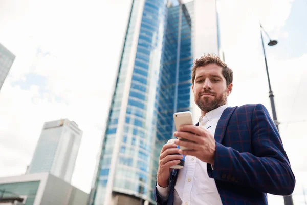 携帯電話でテキスト メッセージを送信して バック グラウンドでオフィス タワーとコーヒーを飲んで市に立っている青年実業家 — ストック写真