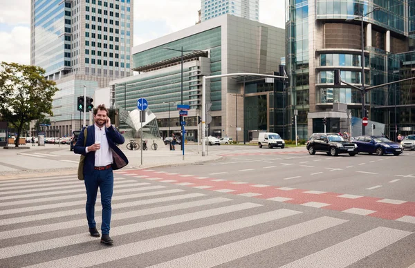 成功的年轻生意人穿西装夹克微笑和谈话在手机上 当横穿街道在城市 — 图库照片