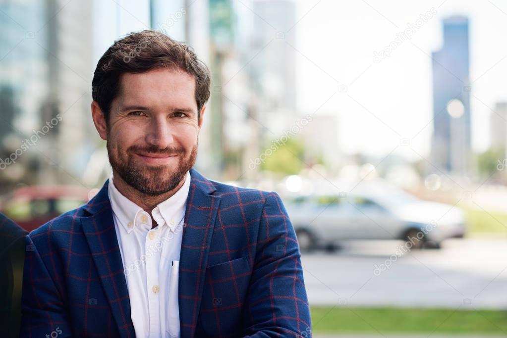 Smiling young businessman wearing blazer standing confidently in the city with office buildings in the background