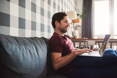 Genç adam tek başına evde onun oturma odasında kanepede otururken dizüstü bilgisayar ile online tarama gülümseyen