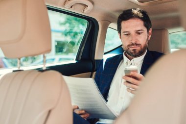 Cep telefonu kullanarak ve şehrin içinden araba araba arka koltuğunda otururken evrak okuma genç işadamı