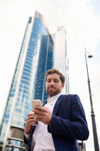 コーヒーを飲むと バック グラウンドでオフィス タワーとの携帯電話のテキスト メッセージを読んで市に立っている青年実業家 — ストック写真