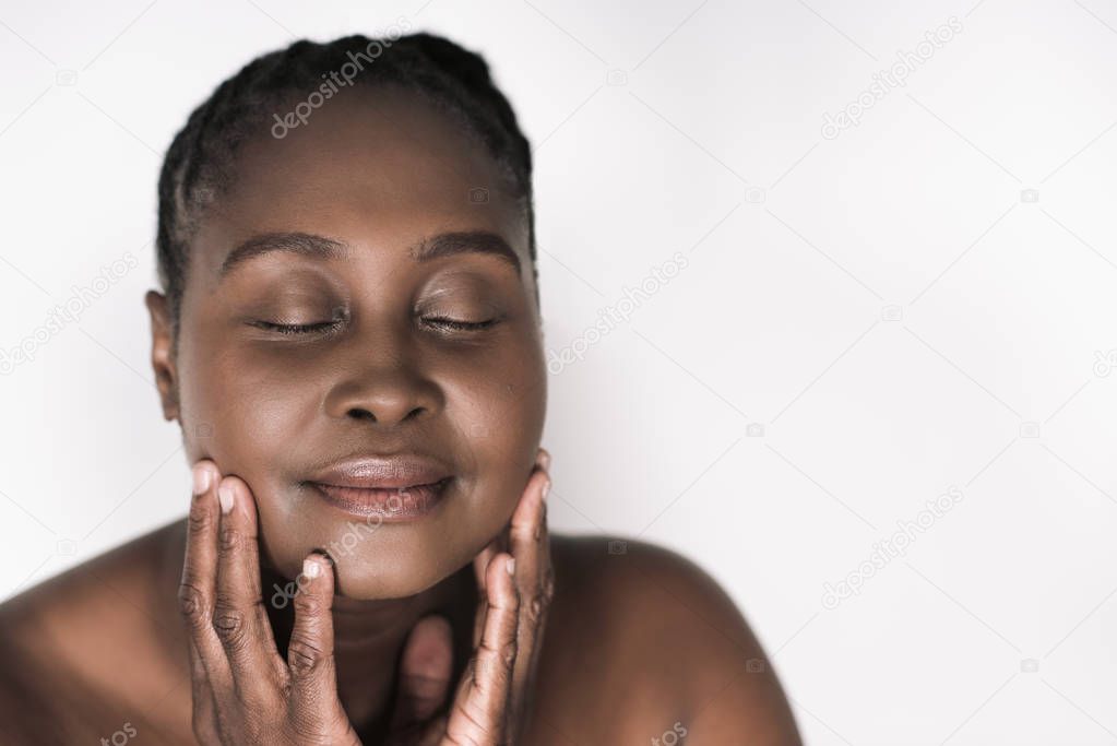 Young plus size African woman with a perfect complexion standing with her eyes closed and touching her face against a white background