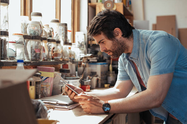 Skilled young craftsman standing at workbench full of tools in his woodworking studio working online with digital tablet