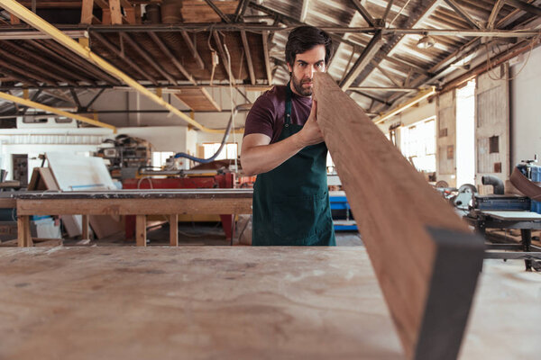 Skilled young woodworker wearing an apron examining the straightness of plank of wood while working alone in his workshop 