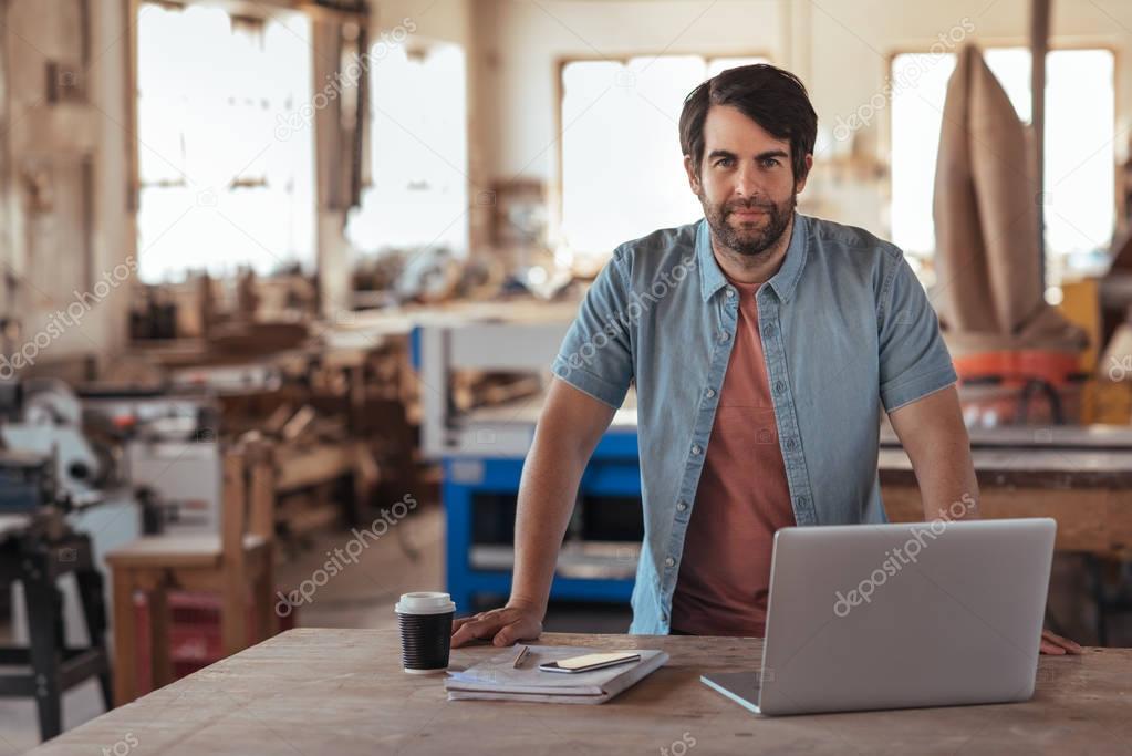 Portrait of a confident young woodworker with beard, leaning over workbench in his large workshop full of carpentry equipment working online with laptop