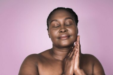Mükemmel bir cilt ile Afrikalı kadın genç artı boyutu, gülümsüyor ve gözleri ile ayakta kapalı ve pembe bir lavanta renkli arka plan yanağında eller