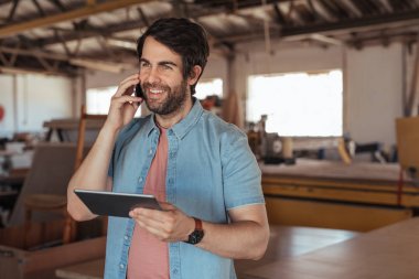 Telefonda konuşmak ve dijital tablet kullanarak büyük doğrama atölyesinde duran sakallı genç woodworker gülümseyen