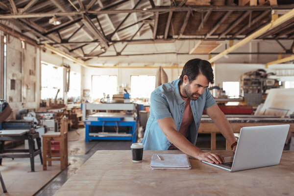 Young woodworker with beard leaning over workbench in his large workshop full of carpentry equipment working online with laptop