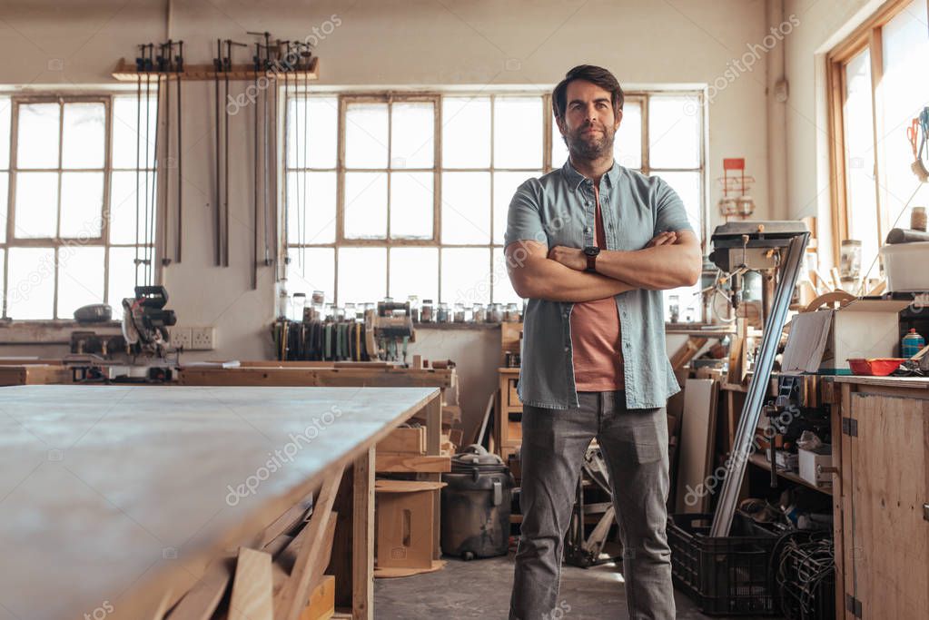 Portrait of skilled young woodworker standing with his arms crossed by workbench full of tools in his carpentry workshop 