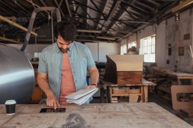 Yetenekli genç woodworker, marangozluk dükkânının dijital tablet kullanma ve tasarımlar defter tutan bir masada duran