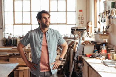 Gülümseyen genç ahşap işçisi, marangozluk atölyesinde alet dolu bir tezgahın yanında dururken yeni tasarımlar düşünüyor