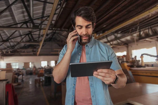 Joven Carpintero Con Barba Hablando Por Celular Usando Una Tableta — Foto de Stock