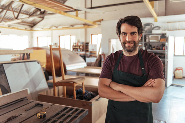 Portrait of a smiling young craftsman standing with his arms crossed by a bench saw in his workshop full of carpentry equipment