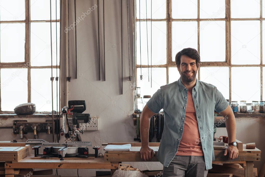 Portrait of a young woodworker smiling confidently while leaning against a workbench full of tools in his carpentry workshop 