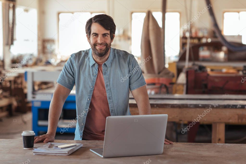Portrait of a smiling young woodworker with a beard, leaning over a workbench in his large workshop full of carpentry equipment working online with a laptop