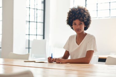 Bir raslantı giyimli genç Afrikalı kız öğrencinin tek başına bir defterde yazılı kampüste bir masada oturan kıvırcık saçlı portresi  
