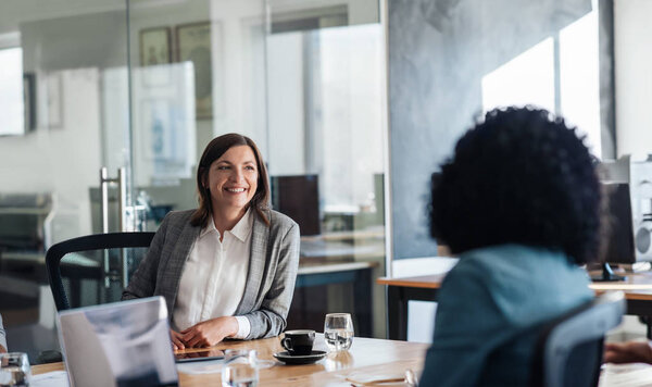 Two smiling young businesswomen talking together while sitting around a boardroom table in a modern office