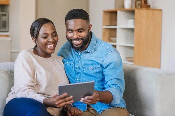 笑着年轻的非洲裔美国夫妇坐在客厅沙发上 用数字平板电脑在网上播放视频 — 图库照片