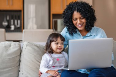 Smiling mother and her cute little daughter watching something together on a laptop while sitting on their living room sofa clipart