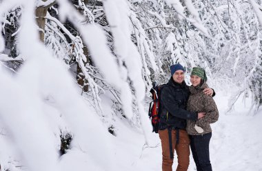 Yürüyüş kıyafetli, karla kaplı bir ormanda, kış yürüyüşü yaparken kol kola duran sevimli genç bir çiftin portresi.