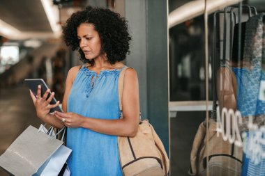 Young woman carrying bags and looking at something on her digital tablet while out shopping for clothes