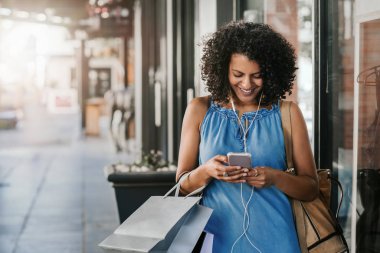 Smiling young woman listening to music on earphones while enjoying a day out shopping for clothing, with flare in the background