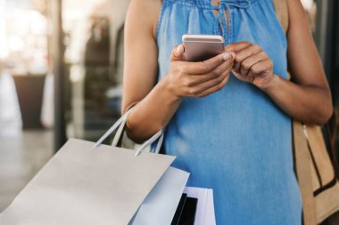 Cropped closeup of a woman carrying bags and sending a text message on her cellphone while out shopping, with flare in the background