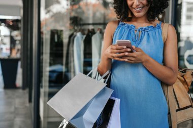 Closeup of a smiling young woman sending a text message on her cellphone while enjoying a day out shopping for clothing