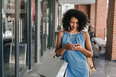 Smiling woman walking down a sidewalk and sending a text on her cellphone while enjoying a day clothes shopping