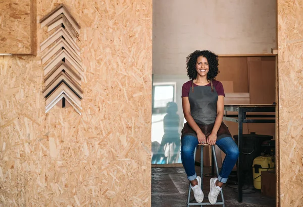 Portrait of a smiling young female entrepreneur wearing an apron sitting on a stool in her picture framing workshop