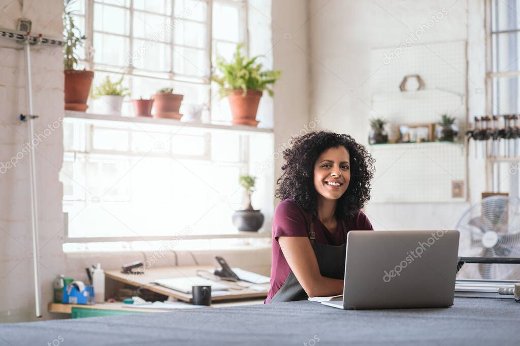 Portrait of a smiling young woman sitting at a workbench in her picture framing studio working on a laptop