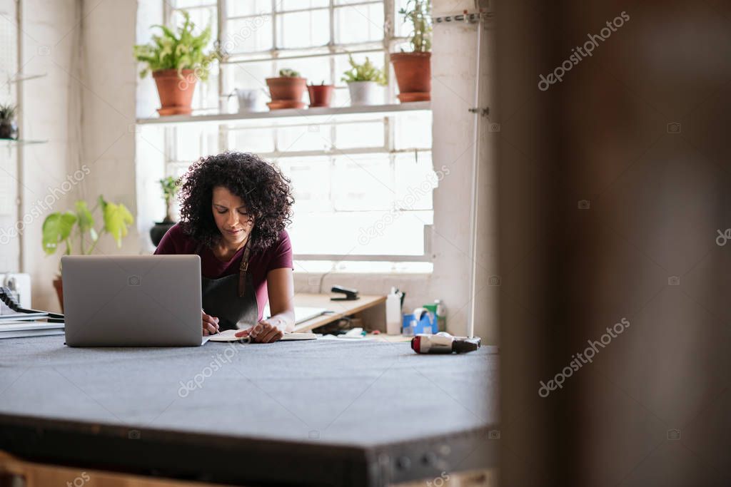 Young woman writing notes and working on a laptop while sitting at a workbench in her picture framing studio