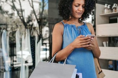 Young woman carrying bags and sending a text message on her cellphone while out shopping for clothing