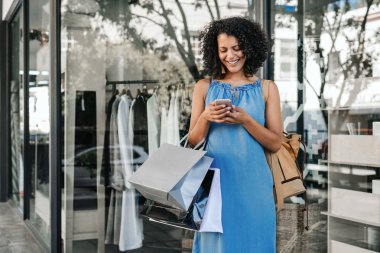 Smiling young woman sending a text message on her cellphone while enjoying a day out shopping for clothing