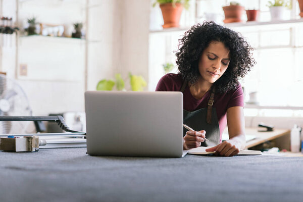 Young woman sitting at a workbench in her picture framing studio writing notes and working online with a laptop
