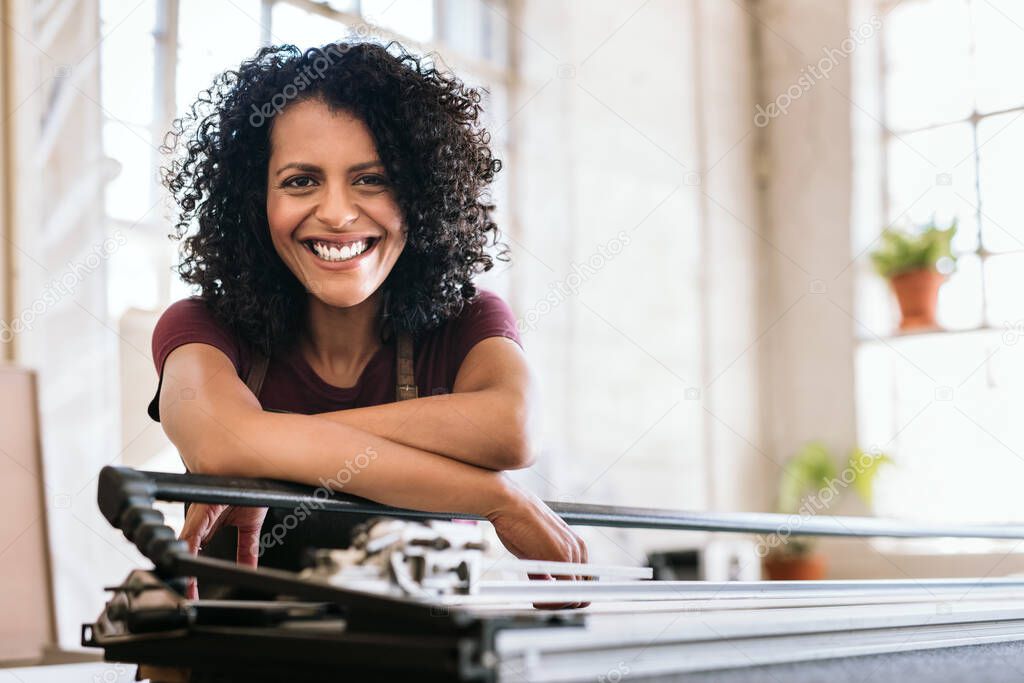 Portrait of a smiling young woman leaning against a mat cutter on a workbench in her picture framing workshop