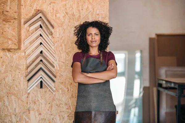 Portrait of a confident young female entrepreneur wearing an apron standing with her arms crossed in her picture framing workshop