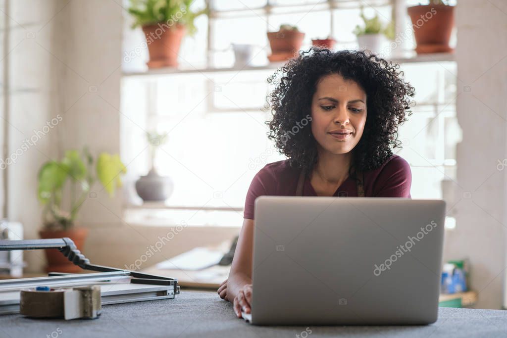 Smiling young woman working on a laptop while sitting at a workbench in her picture framing studio
