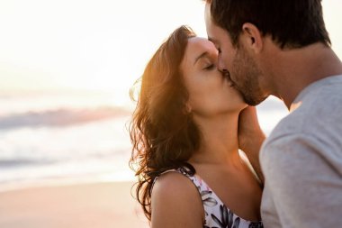 Birbirinin kollarında duran ve alacakaranlıkta kumlu bir sahilde öpüşen genç çift.