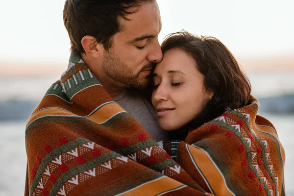 夕暮れ時にビーチの毛布に包まれながら 目を閉じて互いに抱き合う若いカップルを愛情 — ストック写真