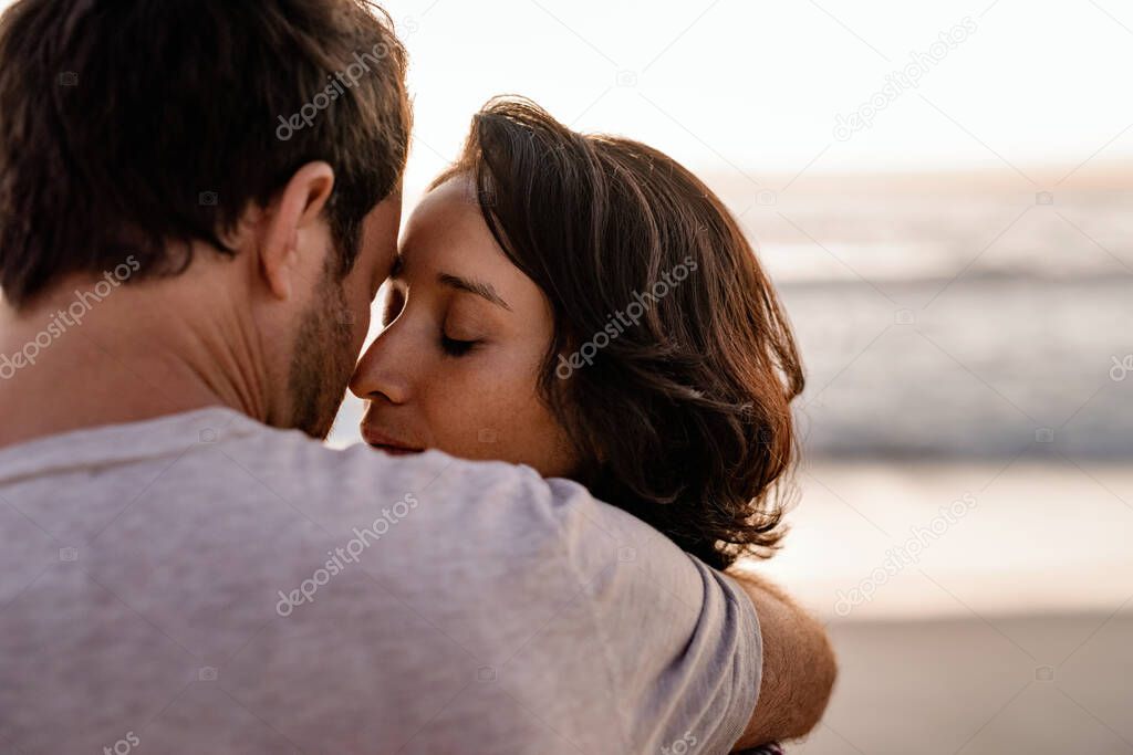 Loving young woman standing with her eyes closed in the arms of her loving husband on a beach at sunset