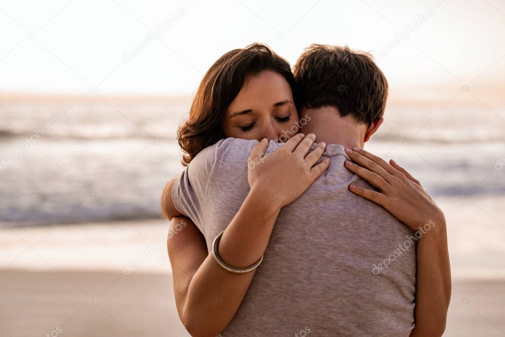 Loving young woman hugging her husband with her eyes closed while standing together on a sandy beach at dusk