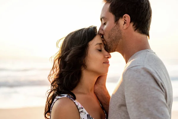 愛情若いです男接吻彼の妻上の彼女の額に一緒に立っている間にビーチに夕暮れ — ストック写真