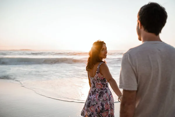 夕日のビーチを歩いている間に海に手で彼女の夫をリード若い女性 — ストック写真