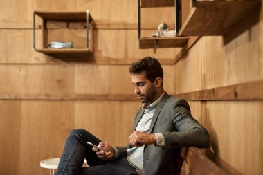 Ceketli şık bir adam modaya uygun bir salonda bankta oturuyor ve cep telefonunu kontrol ediyor.